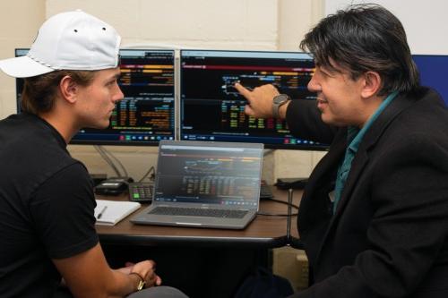 A professor and student sit in front of two computer monitors, each showing financial data